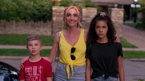 A smiling blond woman in a yellow sleeveless blouse with Ray-bans hanging from the front, on her right her brooding 15 year old biracial daughter in dark colors, on the left her 9 year old blond son in a red tee shirt. They are all taking in the view of their new house (not in image) with varying reactions. 