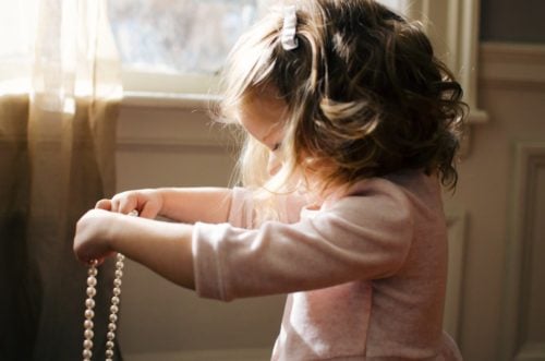 little girl with a hair clip in her curls. She is in profile in a pink shirt and holding out a string of pearls that she is looking down at. There is a curtained window behind her.