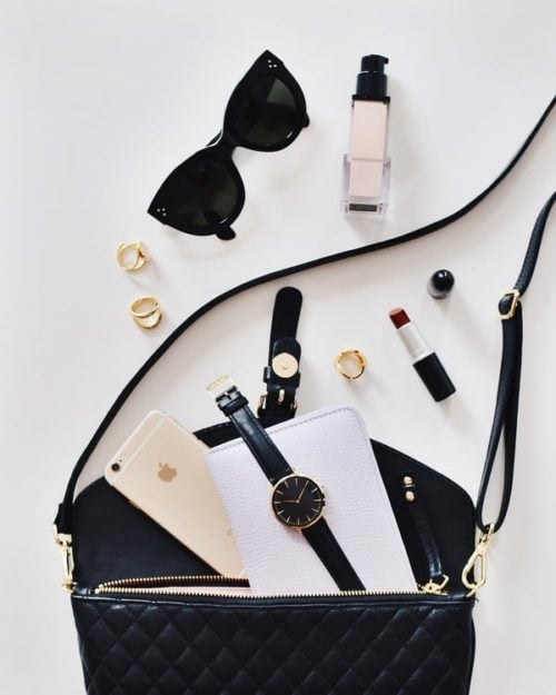 Black handbag on white table, assorted items lay in front of it, sunglasses, lipstick, rings, a watch, and makeup.