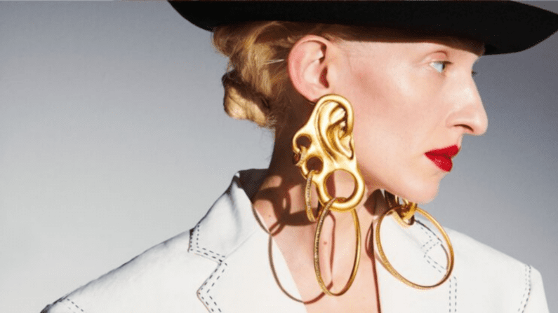 a blond woman in a black hat looks to the side, she is wearing a white shirt and has a bright red lip/ Her giant exaggerated earrings include what looks like a hybrid between a golden ear and brass knuckles with giant hoops hanging from them.